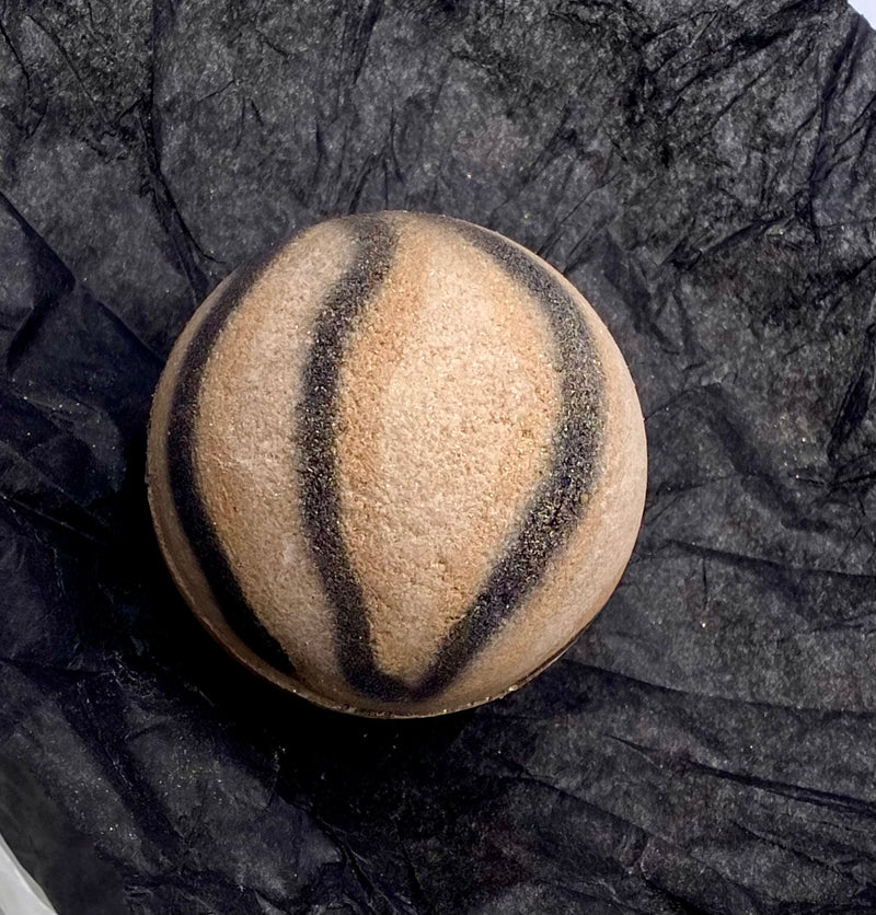 shimmery sand colored round bath bomb with color stripes that mimic tiger stripes of black and gold airbrushed on it. 